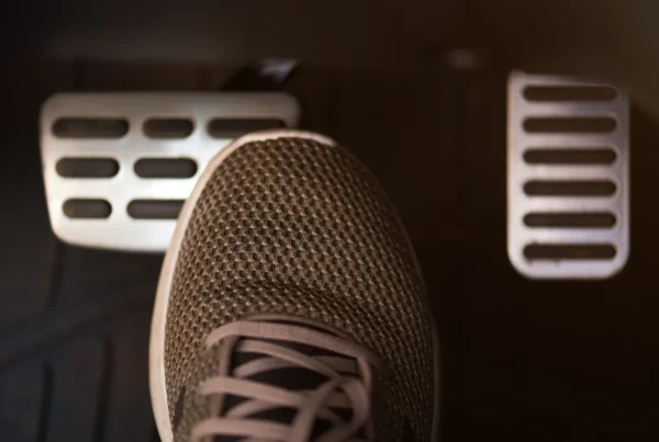 A person's sneaker pressing the brake pedal inside a car's dimly lit footwell, with the accelerator pedal visible to the right.