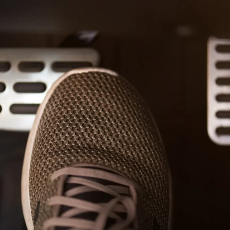 A person's sneaker pressing the brake pedal inside a car's dimly lit footwell, with the accelerator pedal visible to the right.