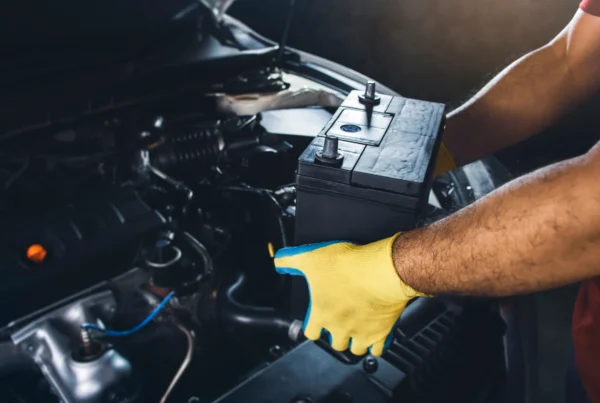 A mechanic wearing a yellow work glove lifts a black car battery out of a vehicle's open engine bay.