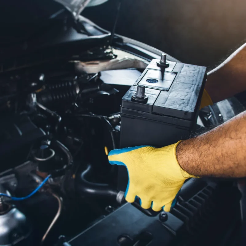 A mechanic wearing a yellow work glove lifts a black car battery out of a vehicle's open engine bay.