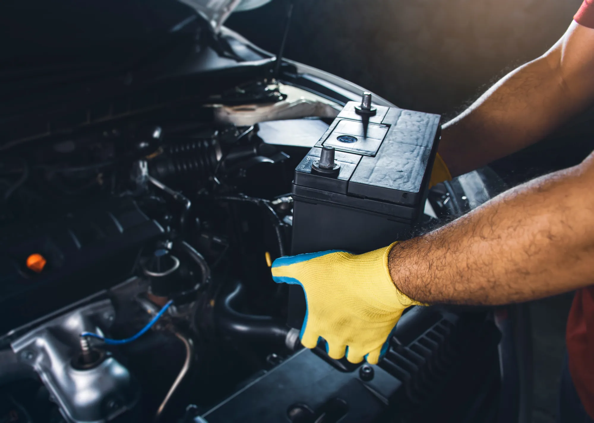 A mechanic wearing a yellow work glove lifts a black car battery out of a vehicle's open engine bay.