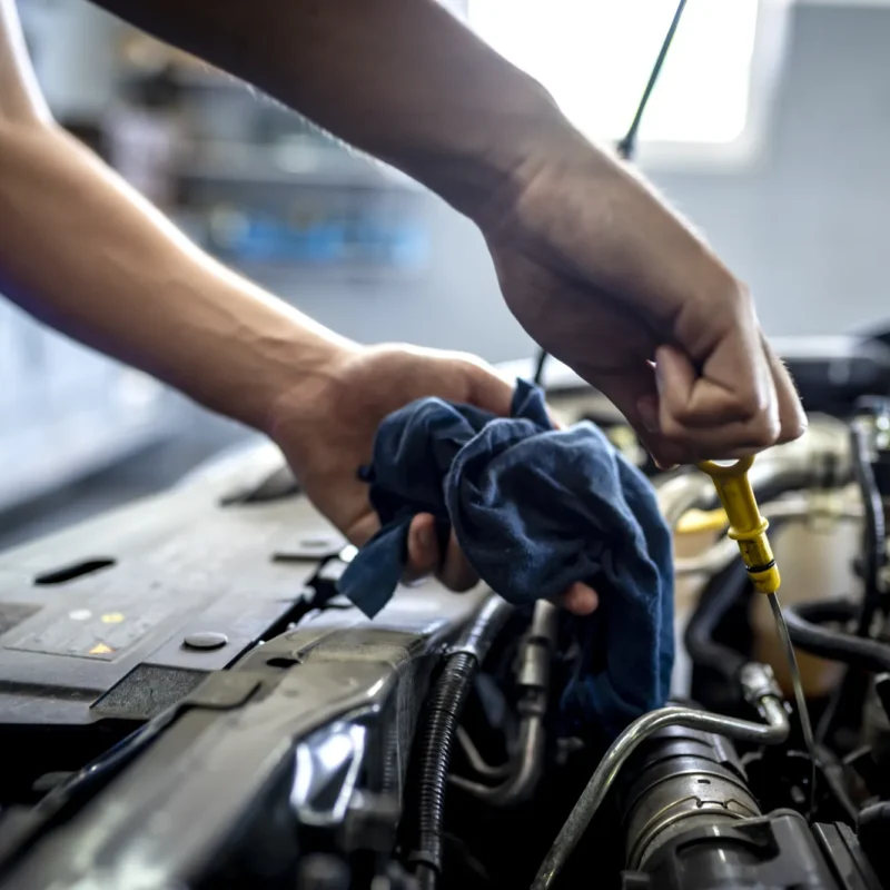 A mechanic in blue coveralls pulls the yellow oil dipstick from a car engine while holding a blue rag.