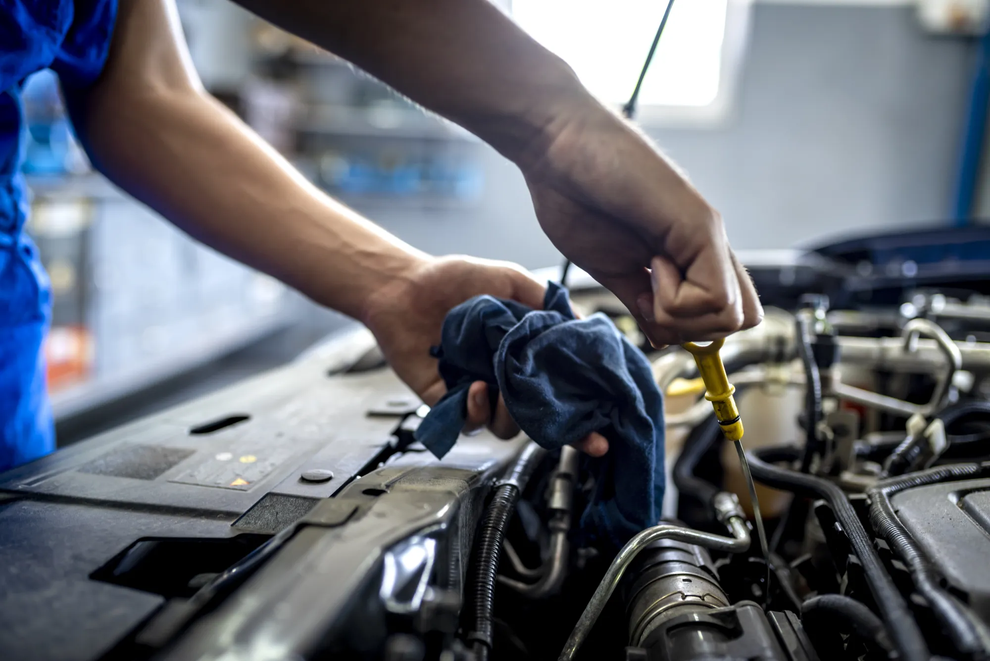 A mechanic in blue coveralls pulls the yellow oil dipstick from a car engine while holding a blue rag.