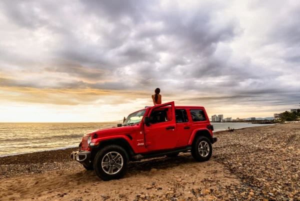 A red four-door Jeep Wrangler parked on a sandy beach at sunset with a woman sitting on its roof facing the ocean.