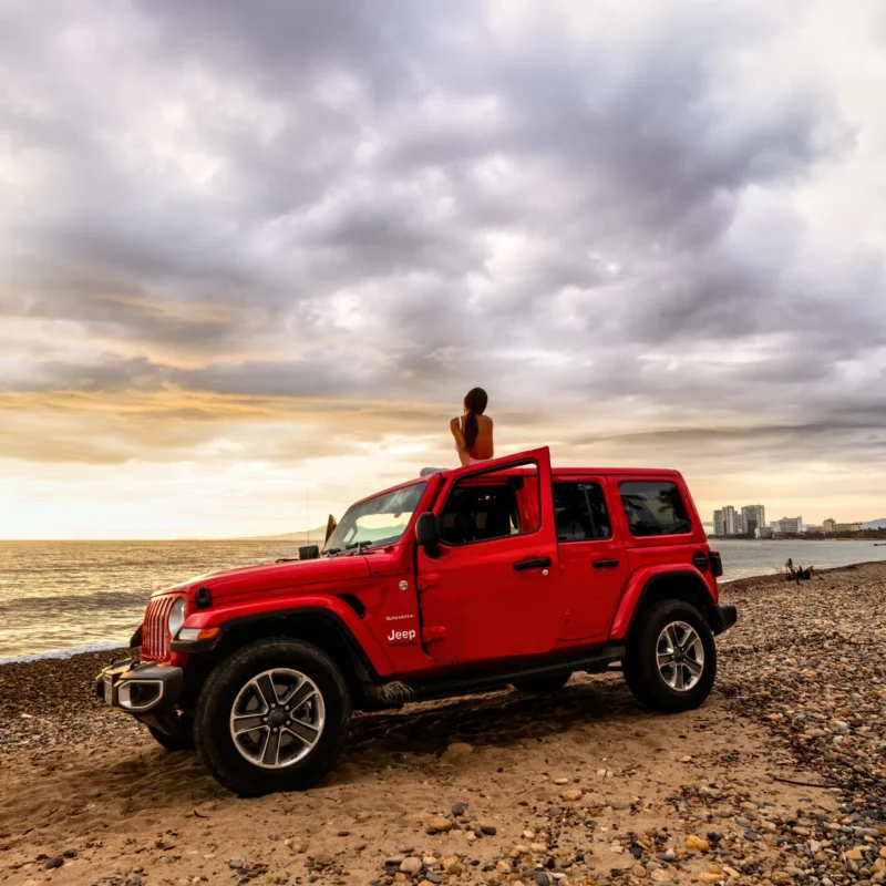 A red four-door Jeep Wrangler parked on a sandy beach at sunset with a woman sitting on its roof facing the ocean.