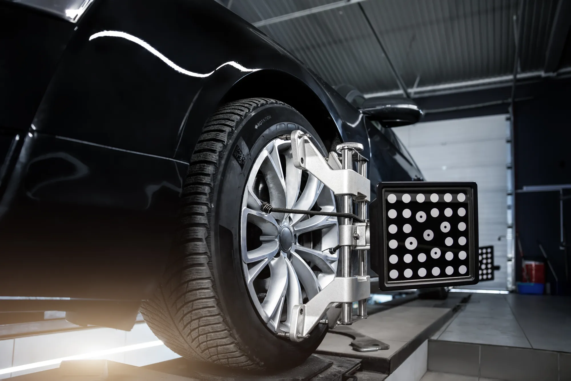 A wheel alignment target board clamped to the alloy wheel of a black car on an alignment rack in a repair shop.