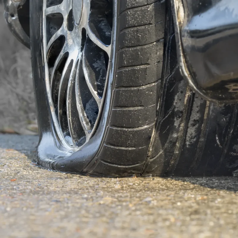 Close-up of a flat, deflated tire on an alloy wheel of a dark car resting on wet asphalt.