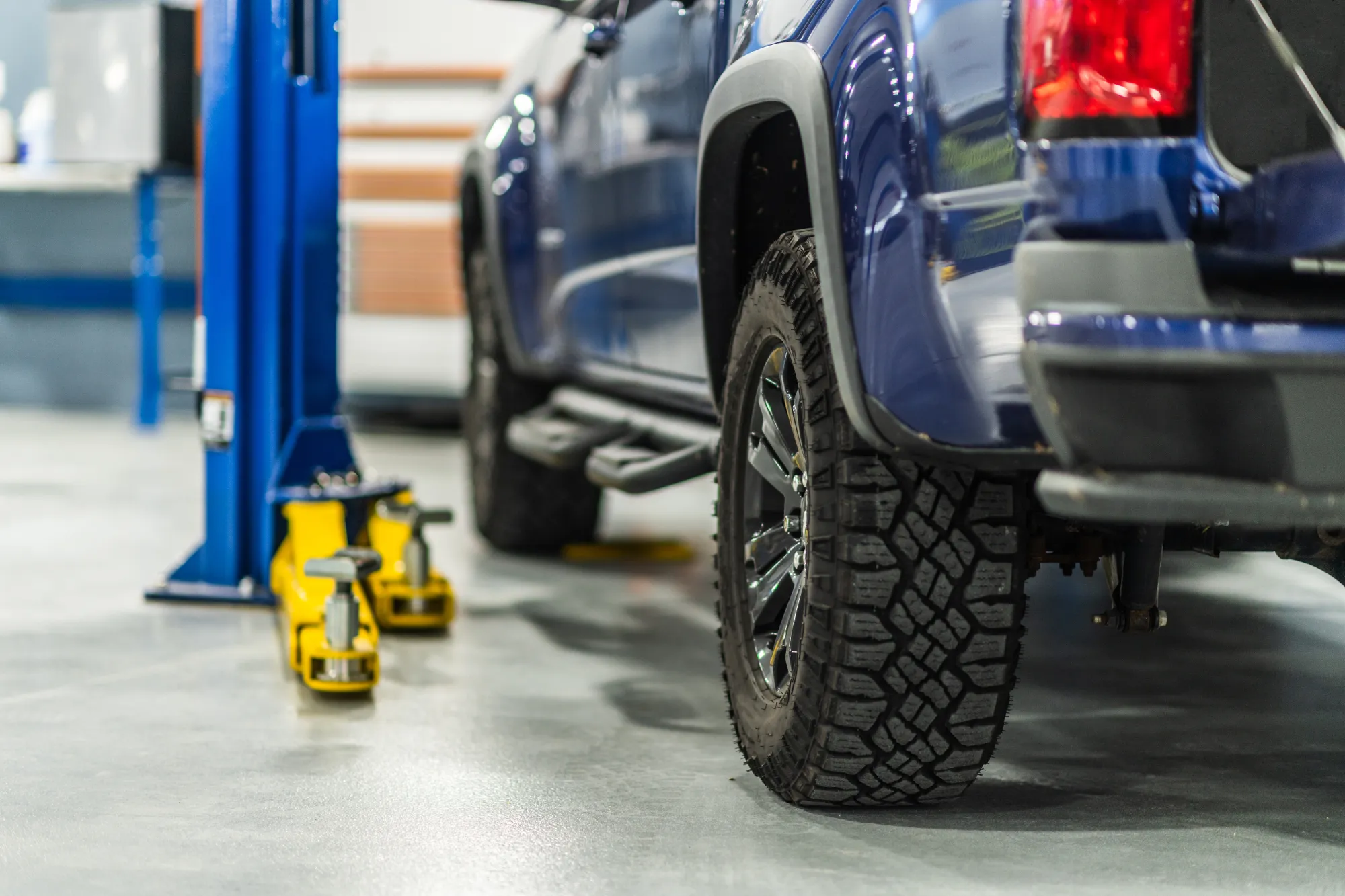 Close-up of the rear all-terrain tire and alloy wheel on a blue pickup truck inside an auto repair shop bay, with a vehicle lift and yellow floor jack in the background.